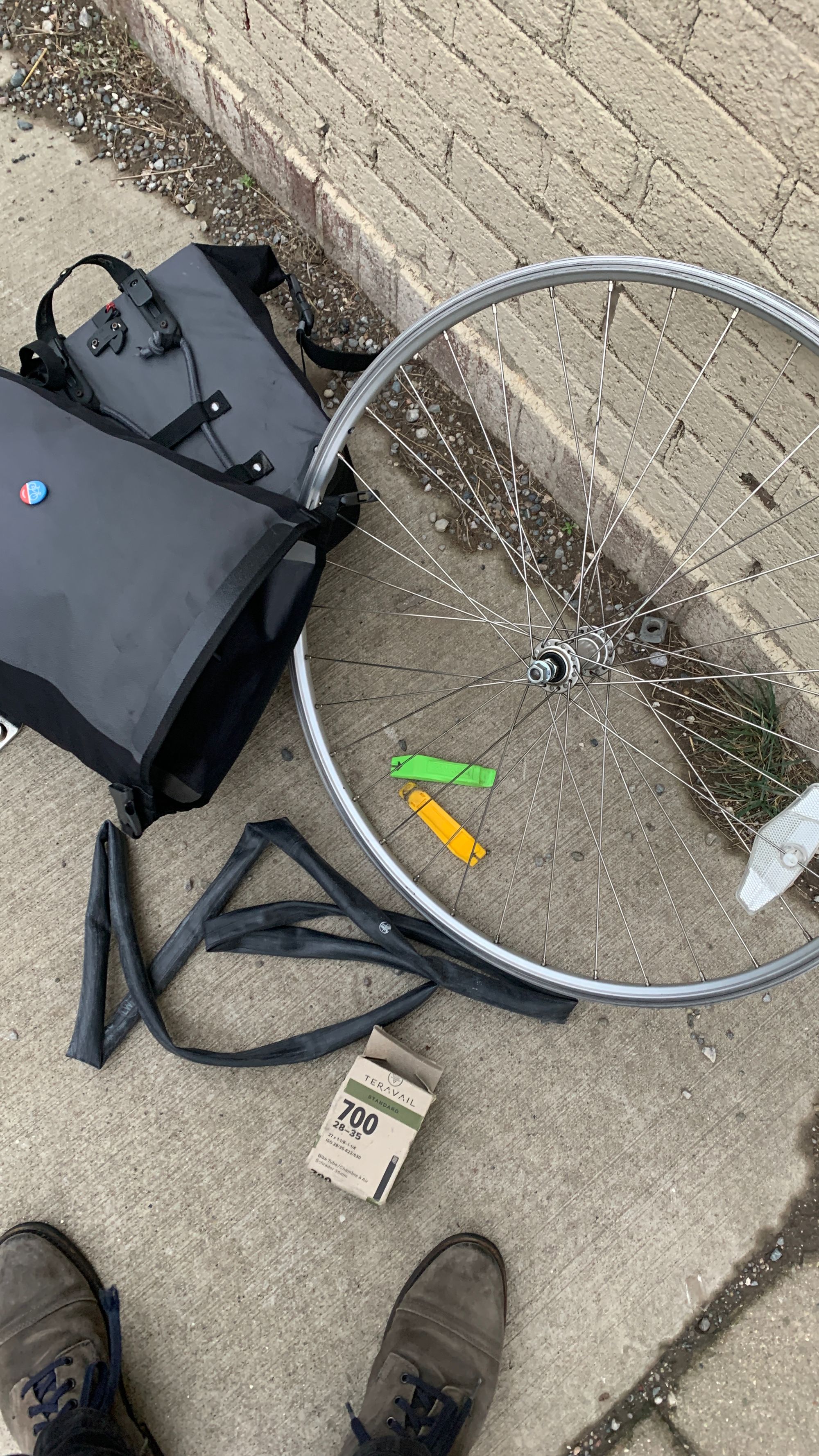 A bicycle wheel on the ground, a punctured inner tube next to the wheel, a pannier bag, tire levers and a spare inner tube. 