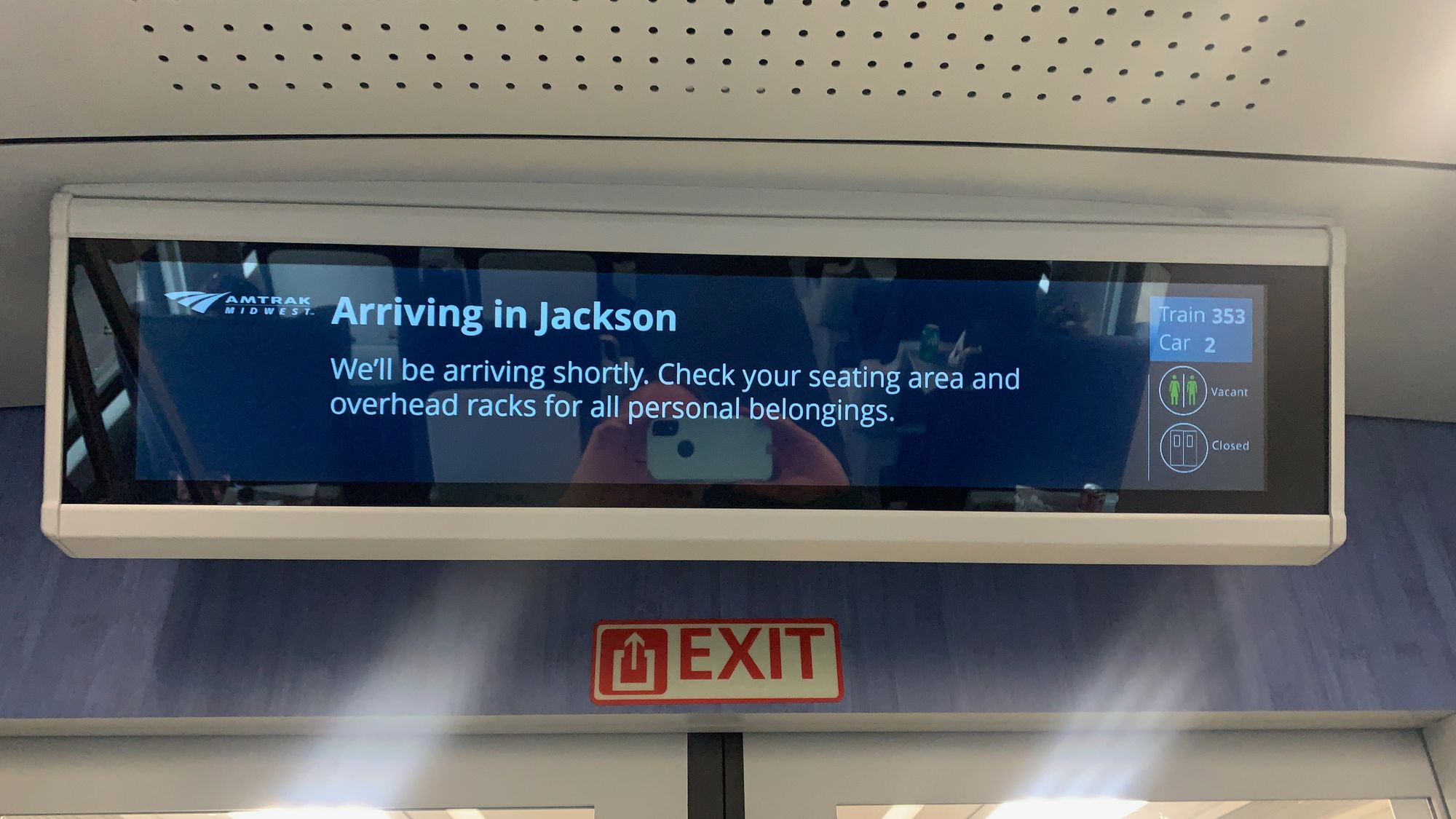 A digital display inside an Amtrak Wolverine coach class car. The screen reads: "Arriving in Jackson. We'll be arriving shortly. Check your seating area and overhead racks for all personal belongings." A sidebar diplay shows the train number, 353, the car number, 2, and shows that the restroom is vacant.